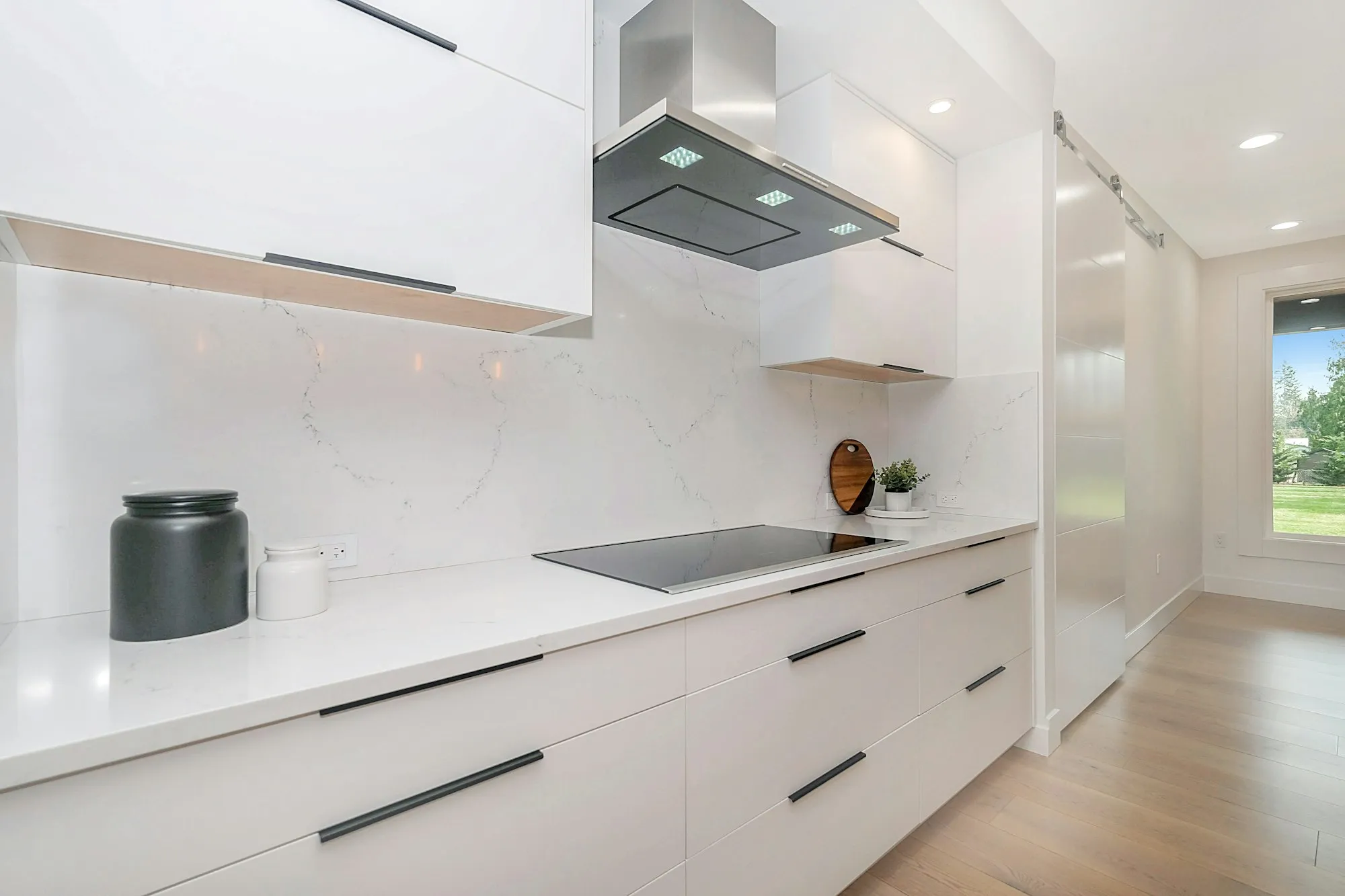 A white handleless kitchen with a marble look splashback, black cooktop and timber floor